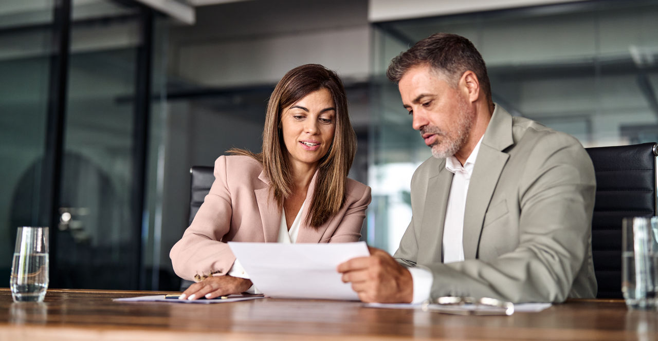 Two professional executives discussing financial accounting papers working together in office. Mature business woman manager consulting older man client holding legal documents at meeting.