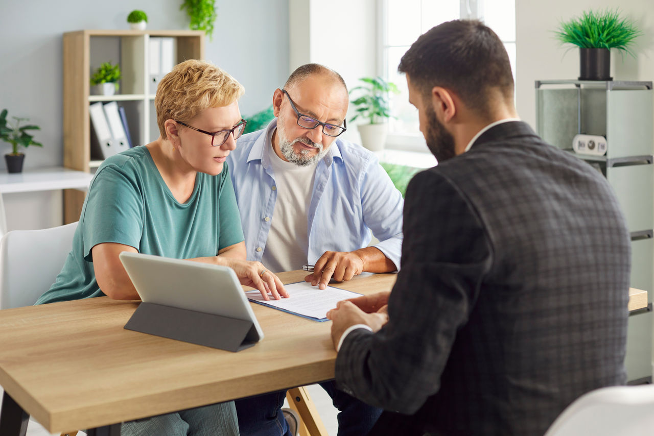 Senior couple sits with advisor reviewing and signing a contract and related document, formalizing agreement during a private meeting at desk. Elderly people make deal with agent in office.